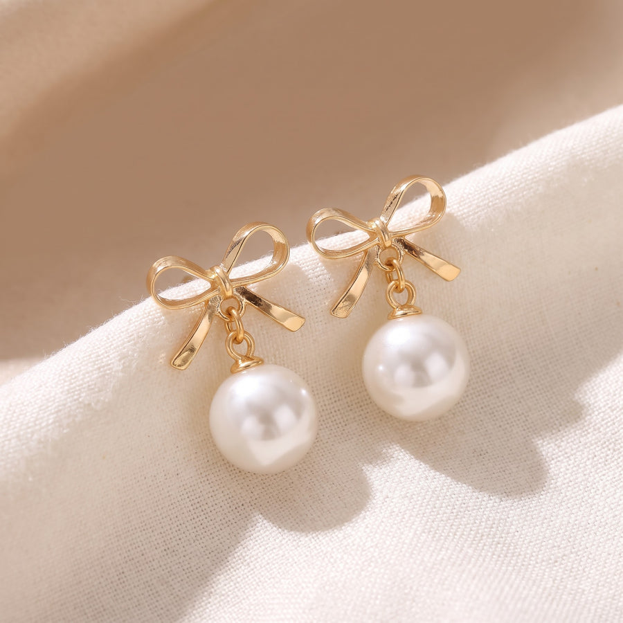 A pair of gold bow stud earrings with white pearl drops, displayed on a soft beige fabric background. - PALLORE Jewellery