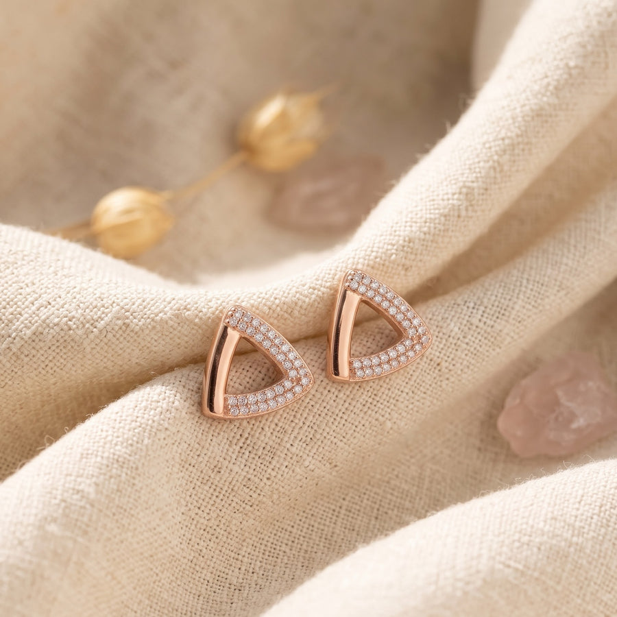 A pair of rose gold triangular stud earrings with pavé-set clear stones, displayed on a textured beige linen fabric with soft decorative stones in the background. - Pallore Jewellery