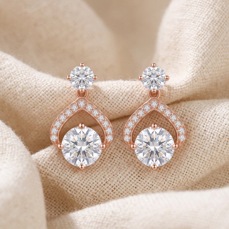 A pair of rose gold drop earrings with large round clear gemstones and smaller accent stones, displayed on a beige textured fabric background. - PALLORE Jewellery