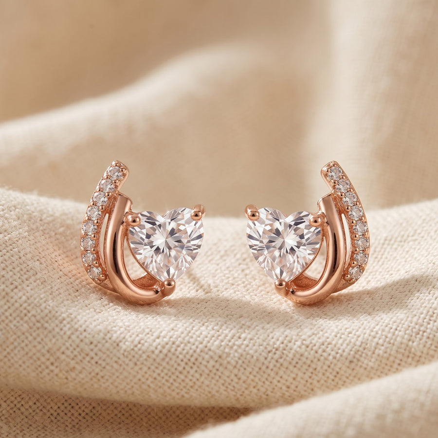 A pair of rose gold crescent-shaped stud earrings with heart-shaped clear gemstones and pavé accents, displayed on a textured beige fabric background. - PALLORE Jewellery
