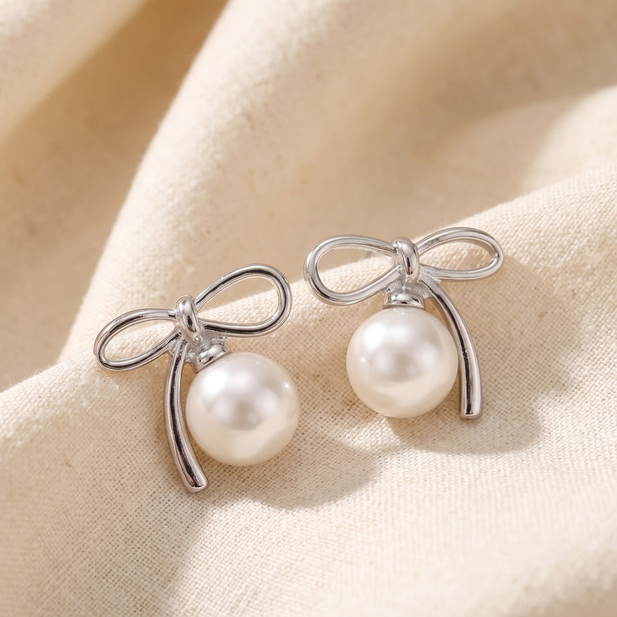 A pair of silver bow stud earrings with round white freshwater pearls, displayed on a soft beige fabric background - PALLORE Jewellery